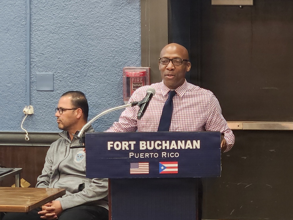 A man is speaking at a podium labeled 'Fort Buchanan, Puerto Rico' with both the American and Puerto Rican flags displayed on the front. He is wearing a pink checkered shirt and dark tie, speaking into a microphone. A second man is seated in the background, wearing glasses and a gray zip-up jacket.