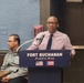 A man is speaking at a podium labeled 'Fort Buchanan, Puerto Rico' with both the American and Puerto Rican flags displayed on the front. He is wearing a pink checkered shirt and dark tie, speaking into a microphone. A second man is seated in the background, wearing glasses and a gray zip-up jacket.