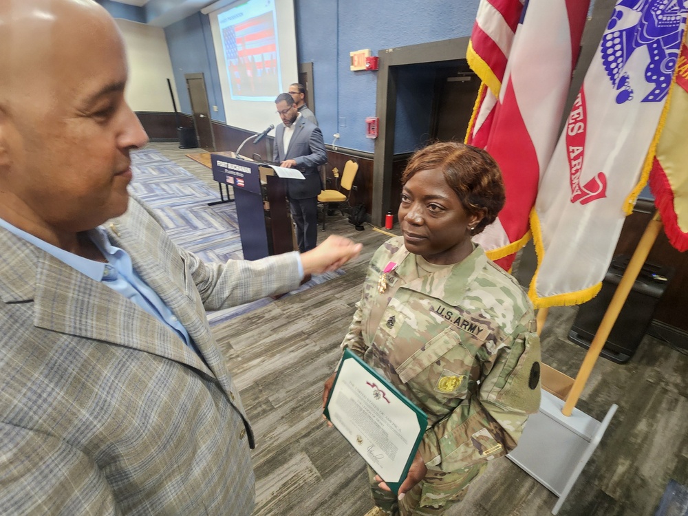 A U.S. Army soldier in OCP (Operational Camouflage Pattern) uniform is receiving recognition at a formal military ceremony, holding a certificate or award document. A civilian in a plaid blazer appears to be congratulating or presenting something to the soldier, who is wearing a medal on their chest. In the background, another individual is speaking at a podium with flags and a projection screen visible.