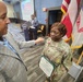 A U.S. Army soldier in OCP (Operational Camouflage Pattern) uniform is receiving recognition at a formal military ceremony, holding a certificate or award document. A civilian in a plaid blazer appears to be congratulating or presenting something to the soldier, who is wearing a medal on their chest. In the background, another individual is speaking at a podium with flags and a projection screen visible.