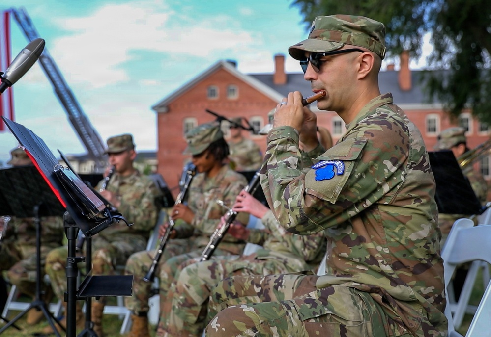 Military personnel in U.S. Army OCP (Operational Camouflage Pattern) uniforms are performing as a military band at an outdoor venue. The foreground soldier is playing a flute or piccolo while wearing sunglasses, with other band members playing clarinets visible behind him. The setting appears to be a formal military installation with a historic brick building visible in the background.