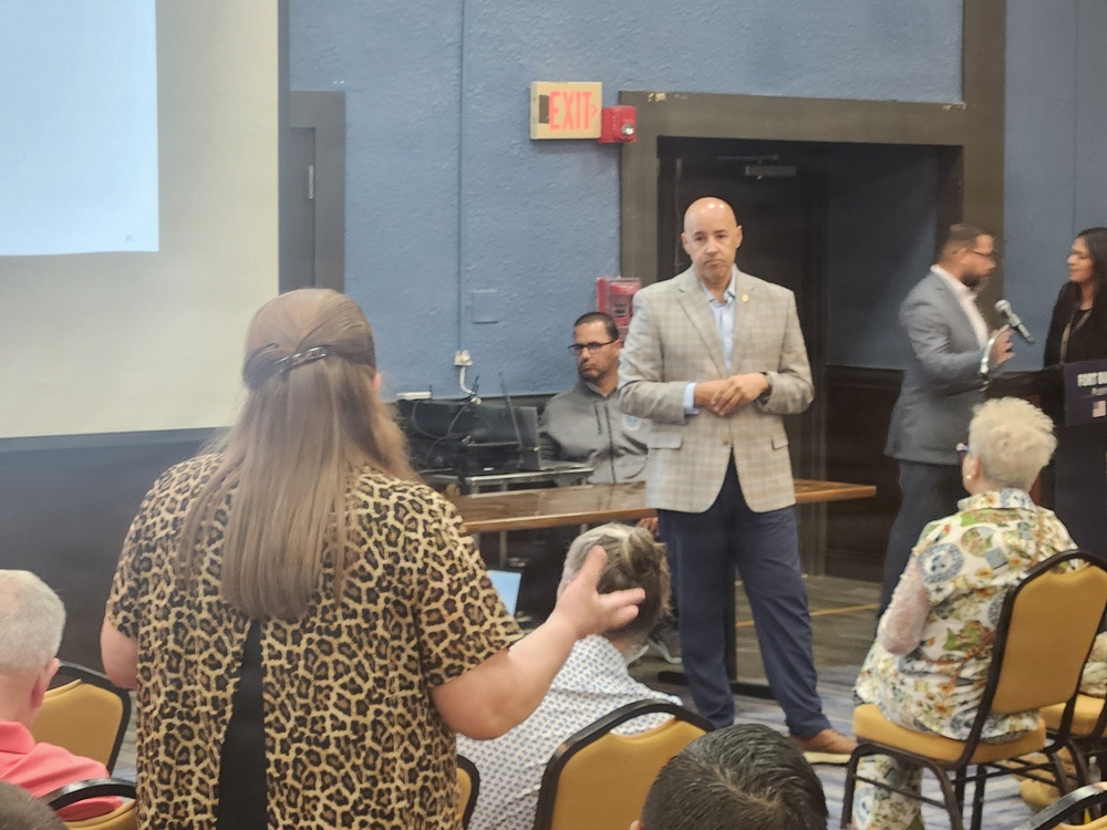 A public meeting or community event is taking place in a conference or meeting room with blue walls. A bald man in a tan plaid blazer and navy pants stands at the front, appearing to address or listen to attendees. Several people are seated in gold/yellow chairs while others stand, with a presentation screen visible on the left and a podium on the right.