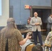 A public meeting or community event is taking place in a conference or meeting room with blue walls. A bald man in a tan plaid blazer and navy pants stands at the front, appearing to address or listen to attendees. Several people are seated in gold/yellow chairs while others stand, with a presentation screen visible on the left and a podium on the right.