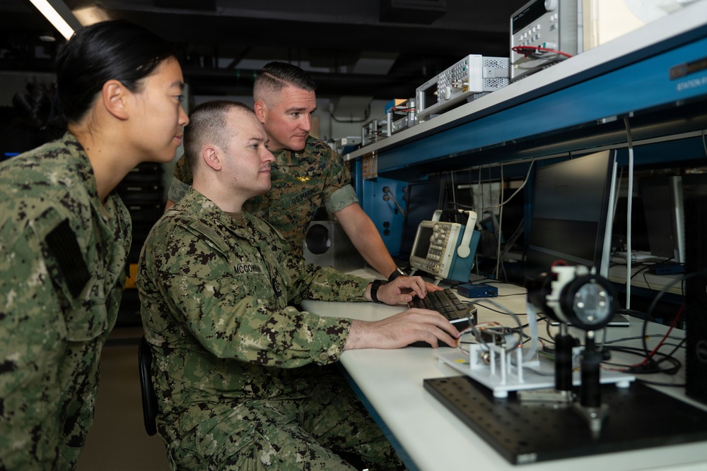 Three military personnel in camouflage uniforms are working together in what appears to be a technical laboratory or electronics workspace. One service member is seated at a workstation operating a keyboard while two others observe. The lab contains various electronic test equipment including an oscilloscope, rack-mounted instruments, and optical components.