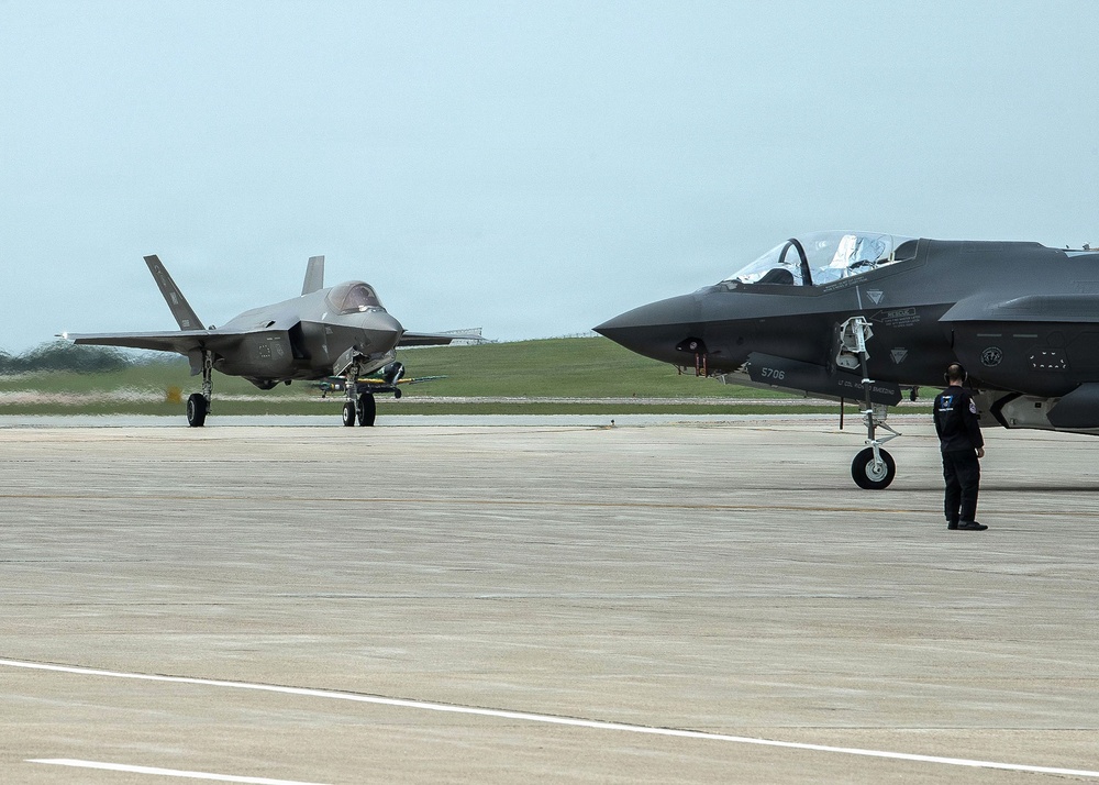 Two F-35 Lightning II stealth fighter jets are visible on a military airfield tarmac. One aircraft is taxiing in the background while another is stationary in the foreground with a ground crew member standing nearby. The scene appears to be a routine flight operations or aircraft marshaling activity.