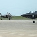 Two F-35 Lightning II stealth fighter jets are visible on a military airfield tarmac. One aircraft is taxiing in the background while another is stationary in the foreground with a ground crew member standing nearby. The scene appears to be a routine flight operations or aircraft marshaling activity.