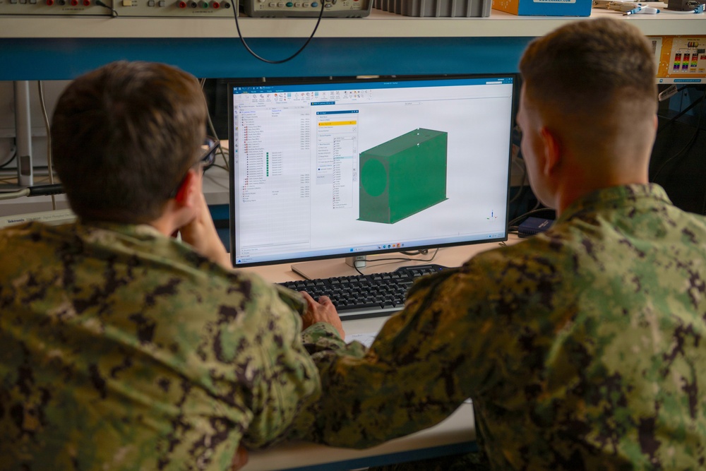 Two military personnel in Navy Working Uniform (NWU) camouflage are reviewing a 3D CAD model on a computer monitor in what appears to be a technical laboratory or engineering workspace. The screen displays a green rectangular box-shaped component in what appears to be CAD software, likely SolidWorks or a similar engineering design application. The setting suggests a military engineering or design review session.