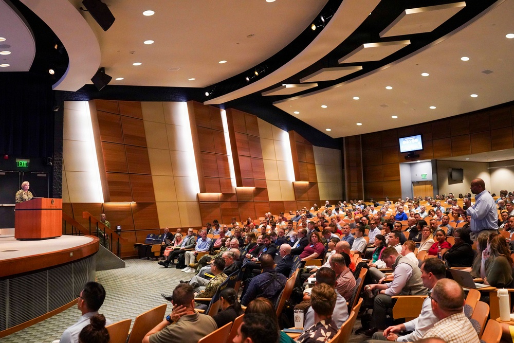 A large auditorium is filled with a diverse audience of several hundred attendees seated in tiered rows of orange/wood-toned chairs. A speaker stands at a podium on a raised stage at the front left, while another individual stands at the right side of the audience. The venue features modern architectural design with wood-paneled walls, recessed lighting, and overhead monitors.
