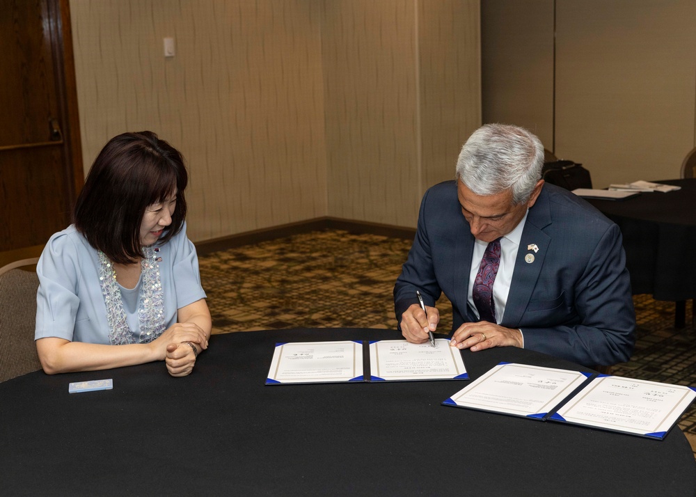 Two individuals are seated at a round table with a black tablecloth, engaged in a formal document signing ceremony. A man in a dark suit is actively signing what appear to be official documents presented in blue folders, while a woman in a light blue dress observes. Multiple copies of the documents are laid out on the table, suggesting a bilateral or institutional agreement.