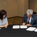 Two individuals are seated at a round table with a black tablecloth, engaged in a formal document signing ceremony. A man in a dark suit is actively signing what appear to be official documents presented in blue folders, while a woman in a light blue dress observes. Multiple copies of the documents are laid out on the table, suggesting a bilateral or institutional agreement.