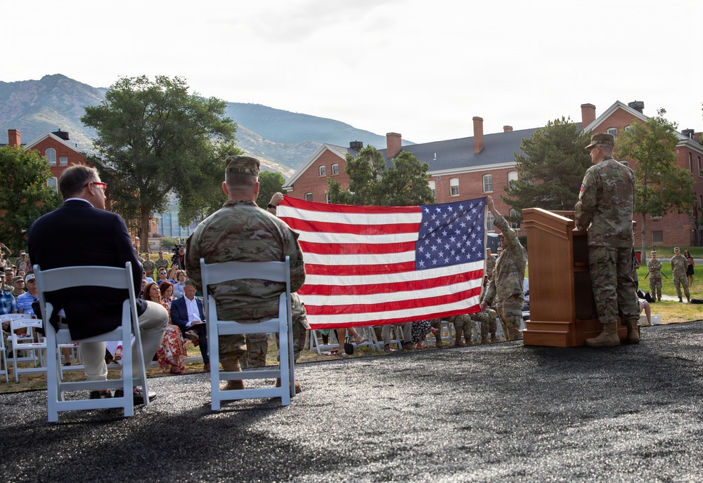 A formal military ceremony is taking place outdoors at what appears to be a military installation, with soldiers in Army Combat Uniforms (ACU/OCP pattern) holding an unfurled American flag. Attendees including civilians and military personnel are seated facing a podium where a soldier appears to be speaking or presiding over the event. The backdrop includes red brick buildings and a mountainous landscape, suggesting a western U.S. military post.