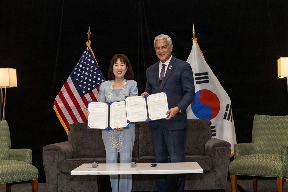 Two individuals are posing together at a formal signing ceremony, each holding an open document with blue covers. The setting features both the American flag and the South Korean flag (Taegukgi) in the background, indicating a bilateral agreement or memorandum of understanding between the United States and South Korea. The event appears to be a professional diplomatic or institutional occasion.