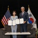 Two individuals are posing together at a formal signing ceremony, each holding an open document with blue covers. The setting features both the American flag and the South Korean flag (Taegukgi) in the background, indicating a bilateral agreement or memorandum of understanding between the United States and South Korea. The event appears to be a professional diplomatic or institutional occasion.