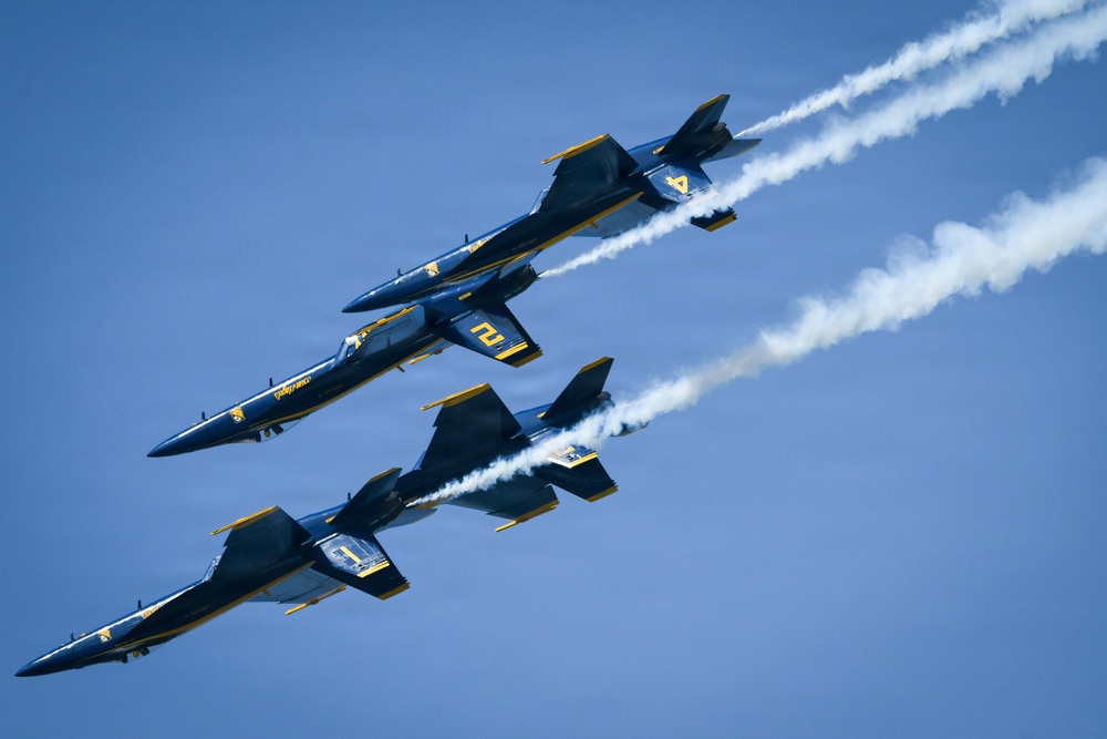 Three U.S. Navy Blue Angels jet aircraft are flying in a tight diagonal formation against a clear blue sky. The aircraft are painted in their distinctive navy blue and gold color scheme and are producing white smoke trails. This appears to be an airshow performance demonstrating precision formation flying.