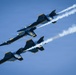 Three U.S. Navy Blue Angels jet aircraft are flying in a tight diagonal formation against a clear blue sky. The aircraft are painted in their distinctive navy blue and gold color scheme and are producing white smoke trails. This appears to be an airshow performance demonstrating precision formation flying.
