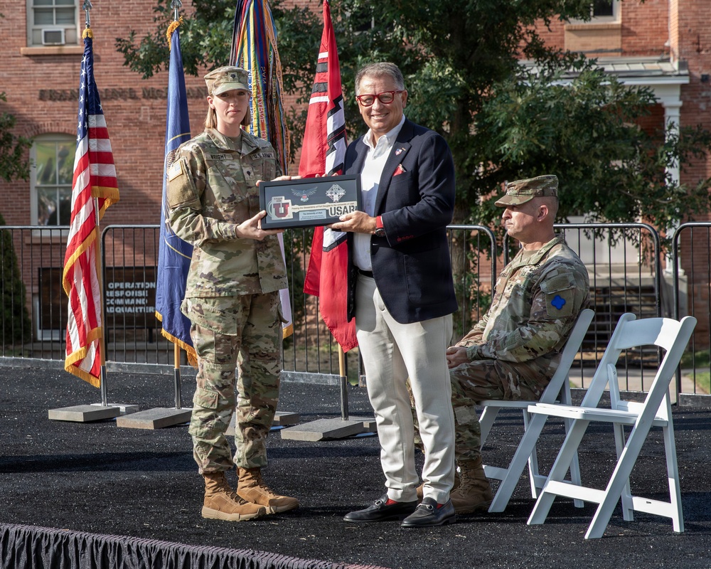 A U.S. Army soldier in multicam uniform is presenting a framed plaque or award to a civilian man in a dark blazer during what appears to be an official military ceremony. The event takes place outdoors on a paved surface in front of a brick building, with military flags and a U.S. flag displayed prominently. A second soldier in uniform is seated in the background observing the presentation.