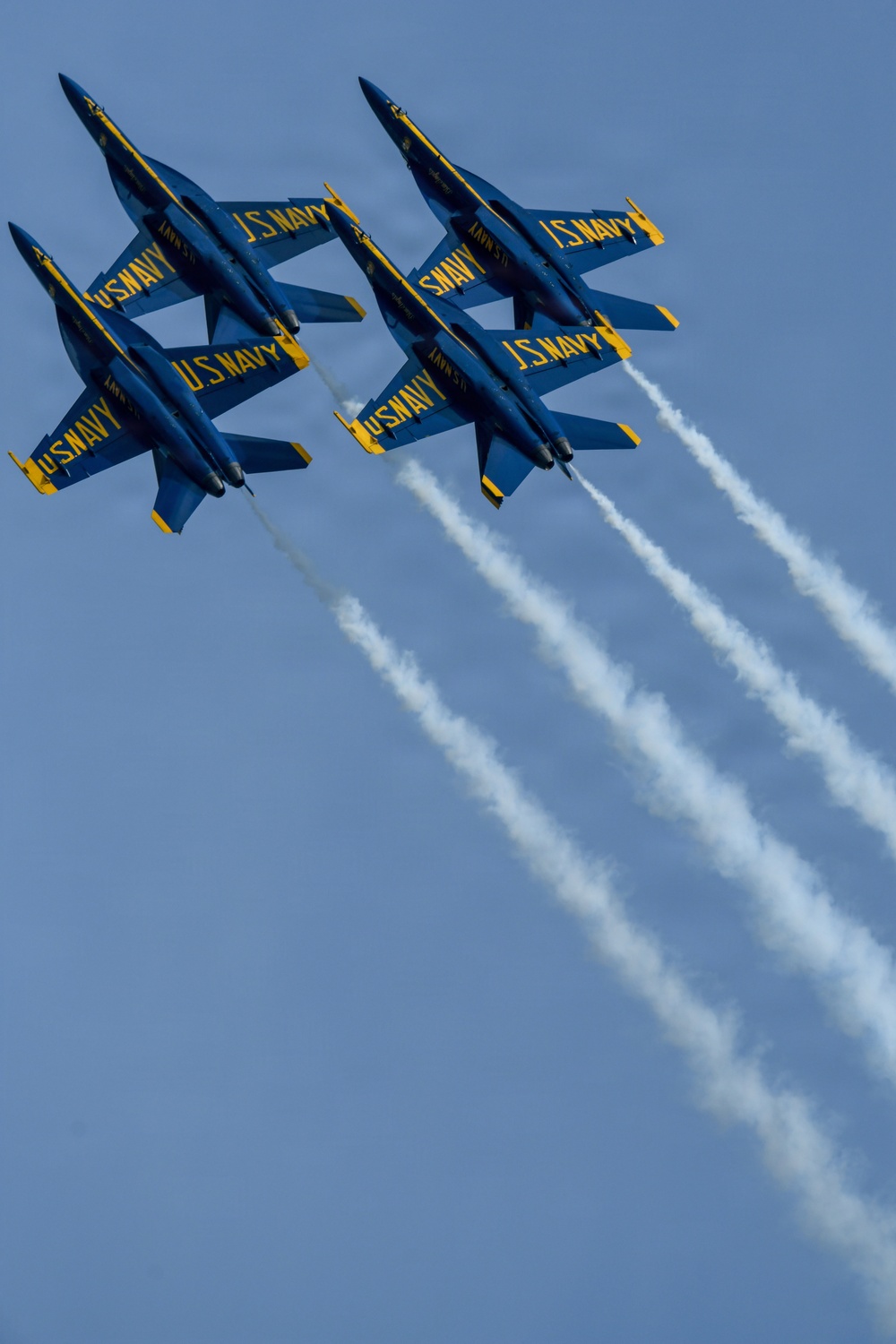Six U.S. Navy Blue Angels F/A-18 Hornet jets are performing a tight formation flight maneuver against a clear blue sky. The aircraft are flying in two groups of three, with several jets appearing inverted, trailing white smoke streams. The image captures a precision aerobatic demonstration characteristic of the Blue Angels flight exhibition team.