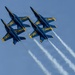 Six U.S. Navy Blue Angels F/A-18 Hornet jets are performing a tight formation flight maneuver against a clear blue sky. The aircraft are flying in two groups of three, with several jets appearing inverted, trailing white smoke streams. The image captures a precision aerobatic demonstration characteristic of the Blue Angels flight exhibition team.