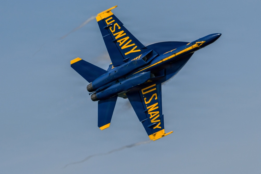 A U.S. Navy Blue Angels F/A-18 Hornet jet is captured in a steep banking maneuver during an airshow performance. The aircraft is painted in the iconic blue and gold color scheme with 'U.S. NAVY' lettering visible on both the top and bottom fuselage. Vapor trails are visible streaming from the wingtips, indicating high-speed, high-G flight conditions.