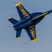 A U.S. Navy Blue Angels F/A-18 Hornet jet is captured in a steep banking maneuver during an airshow performance. The aircraft is painted in the iconic blue and gold color scheme with 'U.S. NAVY' lettering visible on both the top and bottom fuselage. Vapor trails are visible streaming from the wingtips, indicating high-speed, high-G flight conditions.