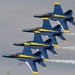 Four U.S. Navy Blue Angels F/A-18 Hornet jets fly in tight diamond formation during an airshow performance. The aircraft display their distinctive blue and yellow livery while producing white smoke trails from their engines. A fifth aircraft is faintly visible in the background lower left, and a tree is partially visible in the foreground left.