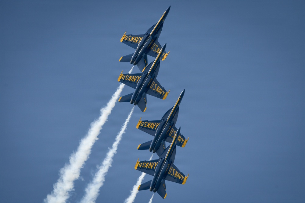Four U.S. Navy Blue Angels F/A-18 Hornet jets fly in tight diamond formation against a clear blue sky. The aircraft are performing an aerobatic maneuver, climbing at a steep angle in close proximity to one another. White smoke trails stream from at least two of the aircraft, enhancing the visual spectacle of the formation flight.