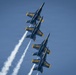 Four U.S. Navy Blue Angels F/A-18 Hornet jets fly in tight diamond formation against a clear blue sky. The aircraft are performing an aerobatic maneuver, climbing at a steep angle in close proximity to one another. White smoke trails stream from at least two of the aircraft, enhancing the visual spectacle of the formation flight.