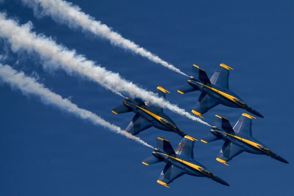 Four military jet aircraft in tight diamond formation fly against a clear deep blue sky, leaving white smoke trails behind them. The aircraft are painted in a distinctive blue and yellow color scheme consistent with the U.S. Navy Blue Angels flight demonstration squadron. The image is captured from a low angle looking up, emphasizing the precision of the formation flying.