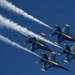 Four military jet aircraft in tight diamond formation fly against a clear deep blue sky, leaving white smoke trails behind them. The aircraft are painted in a distinctive blue and yellow color scheme consistent with the U.S. Navy Blue Angels flight demonstration squadron. The image is captured from a low angle looking up, emphasizing the precision of the formation flying.