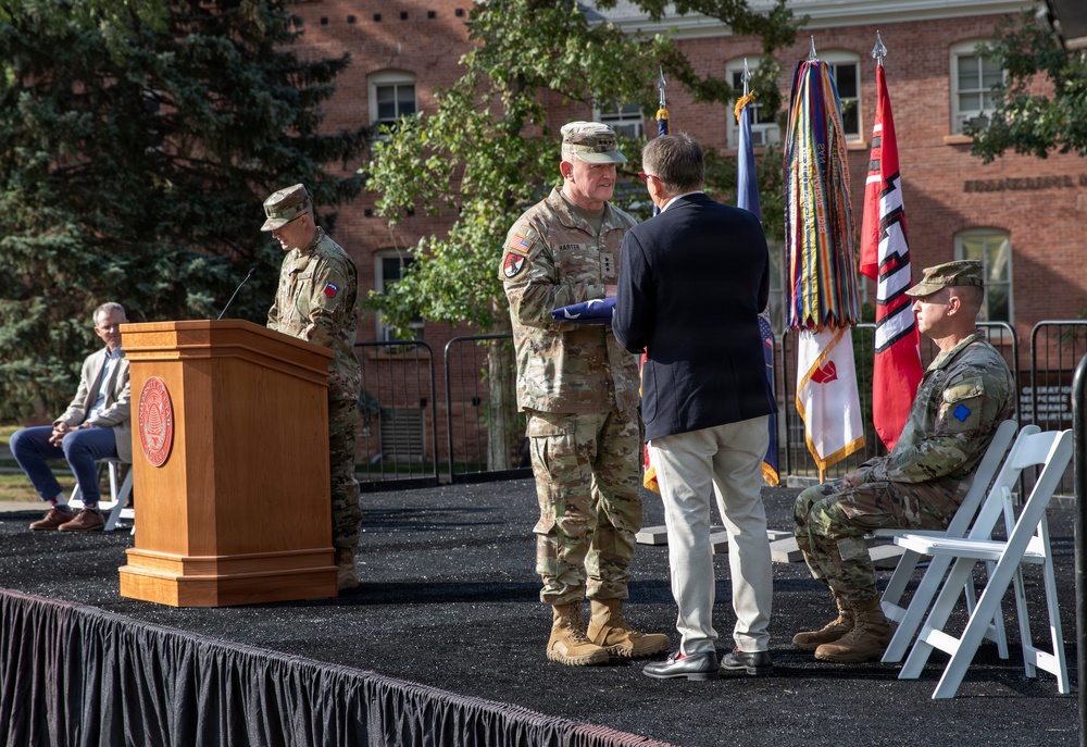 A formal military ceremony is taking place on an outdoor stage in front of a brick building. A soldier in camouflage uniform is presenting or receiving a folded American flag to/from a civilian in a dark blazer, suggesting a retirement, change of command, or memorial ceremony. Additional military personnel and a civilian are present on and near the stage.