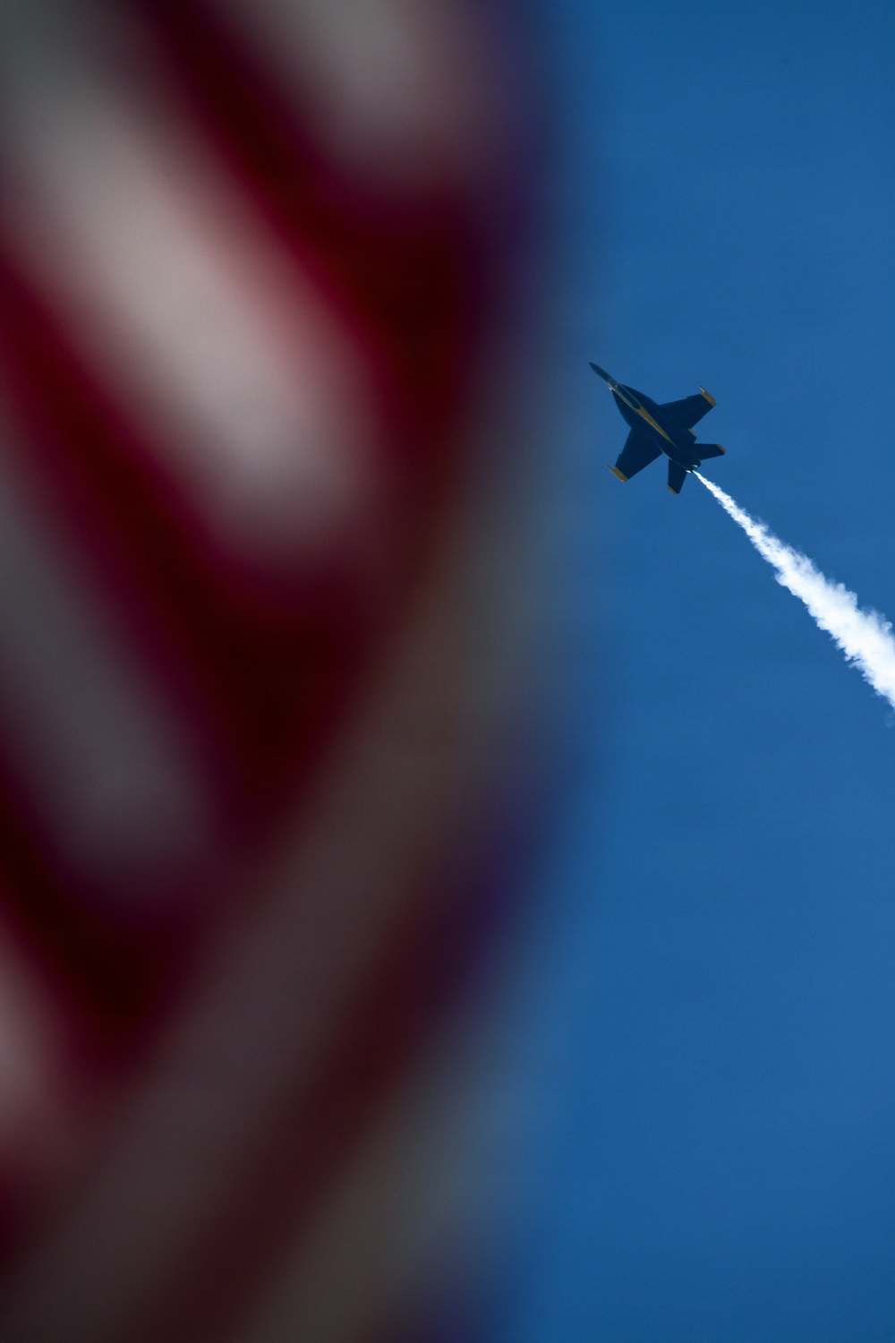 A military jet aircraft, consistent with an F/A-18 Hornet silhouette, is captured climbing steeply through a clear blue sky while leaving a white smoke or contrail trail. An American flag is prominently visible in the foreground, intentionally blurred using shallow depth of field. The composition suggests this is an airshow or patriotic aerial demonstration.