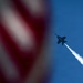 A military jet aircraft, consistent with an F/A-18 Hornet silhouette, is captured climbing steeply through a clear blue sky while leaving a white smoke or contrail trail. An American flag is prominently visible in the foreground, intentionally blurred using shallow depth of field. The composition suggests this is an airshow or patriotic aerial demonstration.
