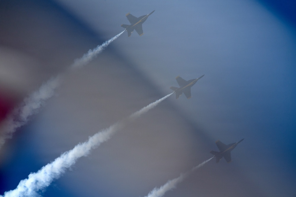 Three military jet aircraft are performing an aerobatic formation flight, leaving white smoke trails behind them as they climb diagonally through a hazy blue sky. The jets appear to be flying in a staggered diagonal formation, consistent with a precision aerobatic display team performance. A blurred foreground element, likely a flag, is partially visible on the left side of the frame.
