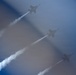 Three military jet aircraft are performing an aerobatic formation flight, leaving white smoke trails behind them as they climb diagonally through a hazy blue sky. The jets appear to be flying in a staggered diagonal formation, consistent with a precision aerobatic display team performance. A blurred foreground element, likely a flag, is partially visible on the left side of the frame.