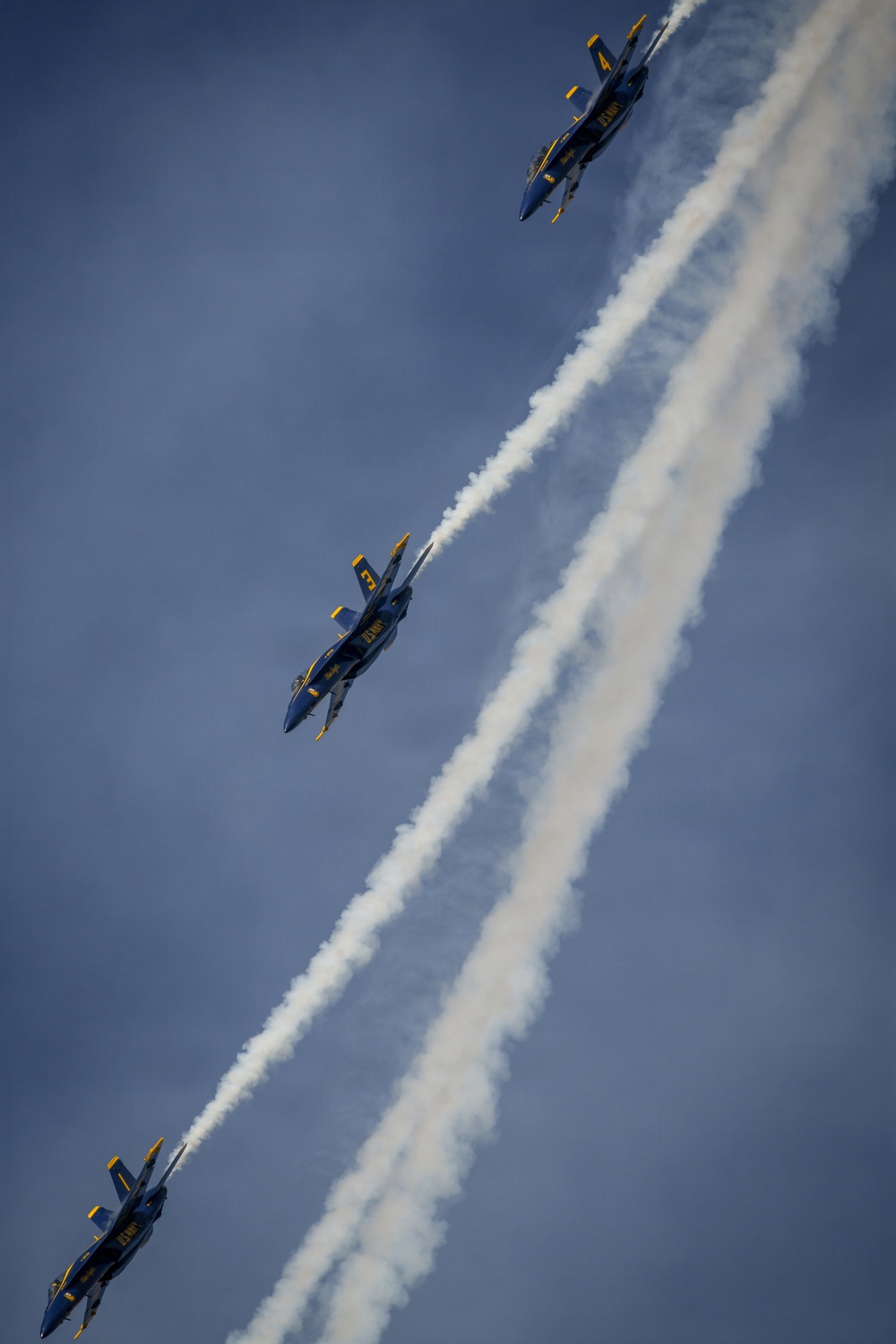 Three U.S. Navy Blue Angels F/A-18 Super Hornet jets are captured in a diagonal echelon formation against a blue-grey sky. Each aircraft is trailing white smoke as they perform a precision aerobatic maneuver. The visible aircraft numbers include #4 (top), #3 (middle), and at least one additional jet at the bottom of the frame.