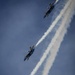 Three U.S. Navy Blue Angels F/A-18 Super Hornet jets are captured in a diagonal echelon formation against a blue-grey sky. Each aircraft is trailing white smoke as they perform a precision aerobatic maneuver. The visible aircraft numbers include #4 (top), #3 (middle), and at least one additional jet at the bottom of the frame.