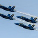 Four U.S. Navy Blue Angels F/A-18 Hornet jets fly in a tight diamond formation against a clear blue sky. The aircraft are numbered 1 through 4 and display the iconic blue and gold color scheme with 'U.S. NAVY' and 'Blue Angels' markings. Each jet produces a white condensation/smoke trail as they perform a precision formation flight maneuver.
