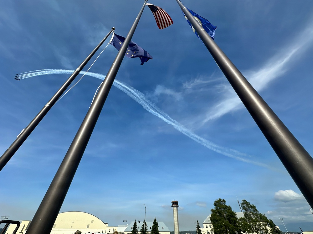 An aerial demonstration team, likely the U.S. Air Force Thunderbirds or Navy Blue Angels, performs a maneuver leaving white smoke trails across a blue sky. The scene is photographed from a low angle looking upward through a cluster of tall flagpoles flying the American flag, the Alaska state flag, and what appears to be a U.S. Air Force flag. A control tower and military base infrastructure are visible in the background, suggesting this is taken at a military air base in Alaska.