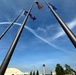 An aerial demonstration team, likely the U.S. Air Force Thunderbirds or Navy Blue Angels, performs a maneuver leaving white smoke trails across a blue sky. The scene is photographed from a low angle looking upward through a cluster of tall flagpoles flying the American flag, the Alaska state flag, and what appears to be a U.S. Air Force flag. A control tower and military base infrastructure are visible in the background, suggesting this is taken at a military air base in Alaska.