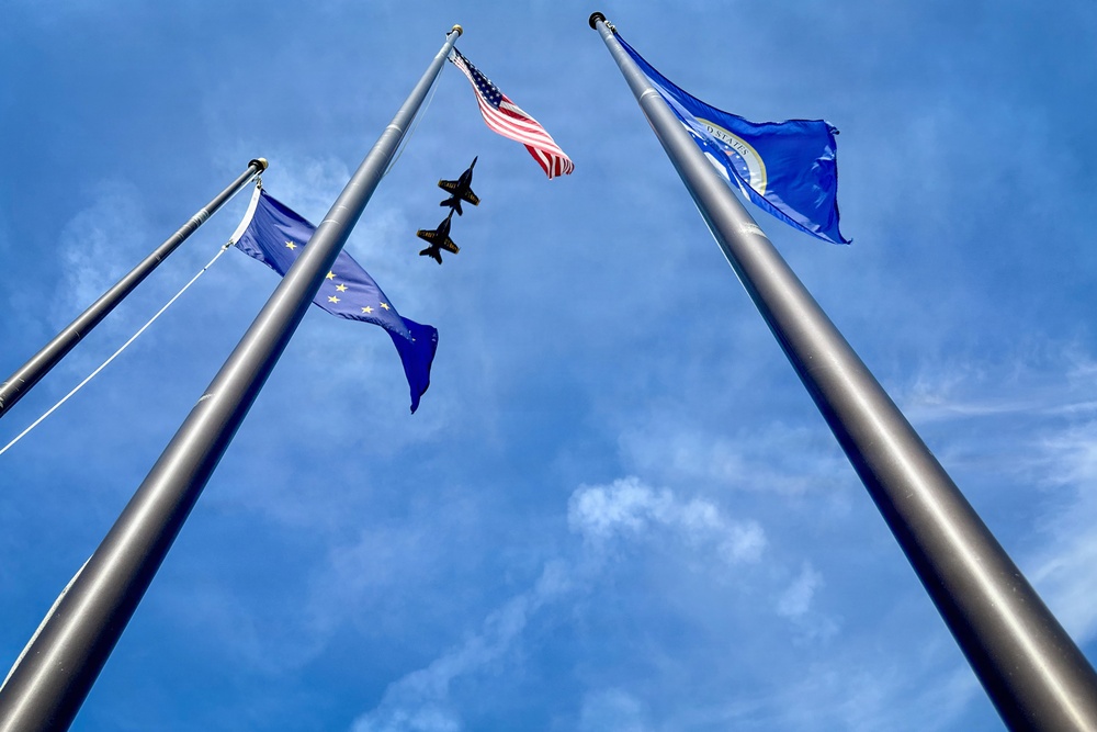 An upward-looking (worm's-eye view) photograph captures three flagpoles against a blue sky with scattered clouds. Three flags are visible — the United States flag, what appears to be the Alaska state flag, and a blue flag consistent with the United States Air Force flag. Two military jet aircraft, likely flying in formation, are visible silhouetted against the sky between the flagpoles.