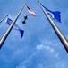 An upward-looking (worm's-eye view) photograph captures three flagpoles against a blue sky with scattered clouds. Three flags are visible — the United States flag, what appears to be the Alaska state flag, and a blue flag consistent with the United States Air Force flag. Two military jet aircraft, likely flying in formation, are visible silhouetted against the sky between the flagpoles.
