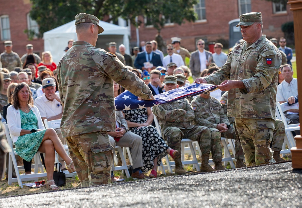 Two U.S. Army soldiers in OCP (Operational Camouflage Pattern) uniforms are performing a flag folding ceremony at an outdoor military event. A large audience of both military personnel and civilians is seated in white chairs observing the ceremony. The setting appears to be on a military installation, with brick buildings visible in the background.