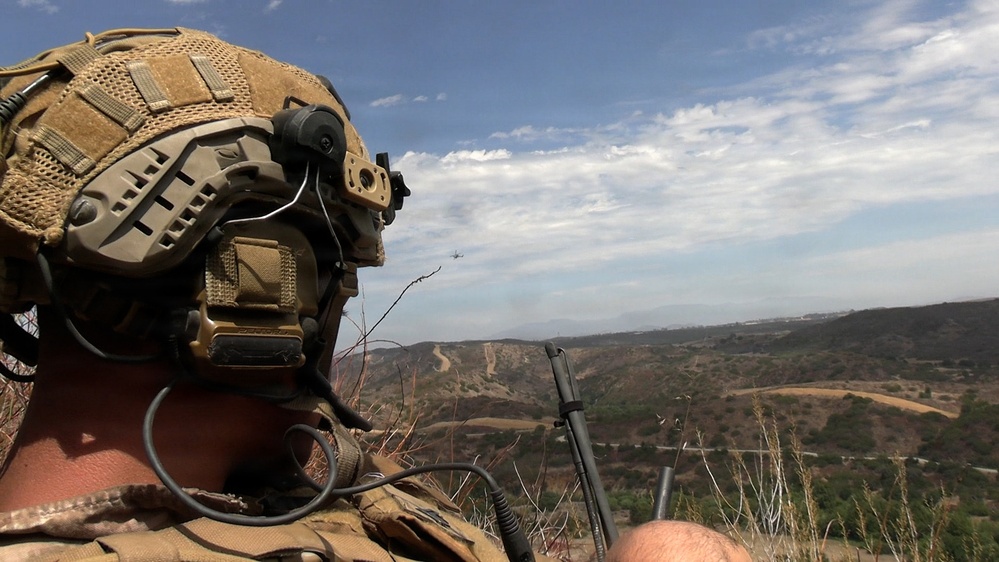 A military or special operations personnel is positioned on an elevated terrain overlooking a dry, hilly landscape. The individual is wearing a tactical helmet with communications equipment and appears to be observing or coordinating with an aircraft visible in the distance. The setting appears to be a training exercise or operational deployment in an arid, semi-rural environment.