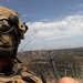 A military or special operations personnel is positioned on an elevated terrain overlooking a dry, hilly landscape. The individual is wearing a tactical helmet with communications equipment and appears to be observing or coordinating with an aircraft visible in the distance. The setting appears to be a training exercise or operational deployment in an arid, semi-rural environment.