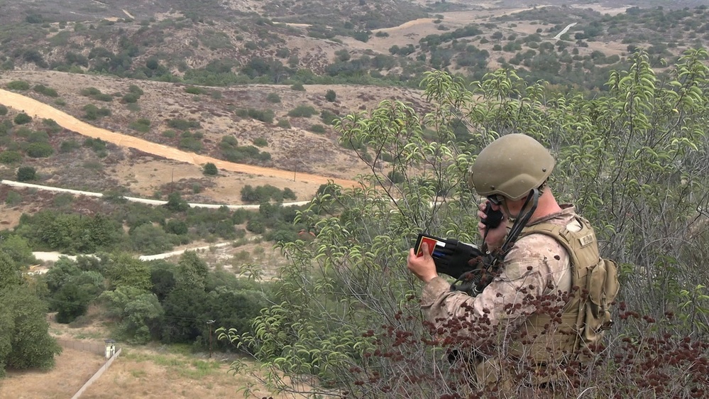 A military service member in desert camouflage uniform and tactical helmet is positioned on an elevated hillside overlooking a valley. The soldier is using a handheld radio/communication device while holding what appears to be a tablet or map reference card. The terrain suggests a training environment, likely a U.S. military installation based on the landscape characteristics.