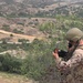 A military service member in desert camouflage uniform and tactical helmet is positioned on an elevated hillside overlooking a valley. The soldier is using a handheld radio/communication device while holding what appears to be a tablet or map reference card. The terrain suggests a training environment, likely a U.S. military installation based on the landscape characteristics.