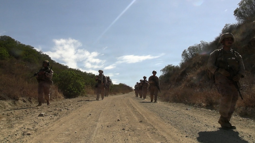 A group of armed military personnel in full combat gear are conducting a patrol along a dirt road flanked by dry, scrubby hillsides. The soldiers are wearing helmets and carrying rifles, moving in a loose column formation toward the camera. The terrain and vegetation suggest an arid or semi-arid environment, consistent with a training exercise or operational patrol.