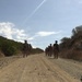 A group of armed military personnel in full combat gear are conducting a patrol along a dirt road flanked by dry, scrubby hillsides. The soldiers are wearing helmets and carrying rifles, moving in a loose column formation toward the camera. The terrain and vegetation suggest an arid or semi-arid environment, consistent with a training exercise or operational patrol.