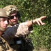 Two military personnel in combat gear are shown in an outdoor setting with dense vegetation in the background. One individual wearing sunglasses and a tactical helmet is pointing in a specific direction, appearing to brief or direct the other soldier. Both are equipped with body armor, helmets, and communication equipment consistent with U.S. Marine Corps desert MARPAT camouflage uniforms.