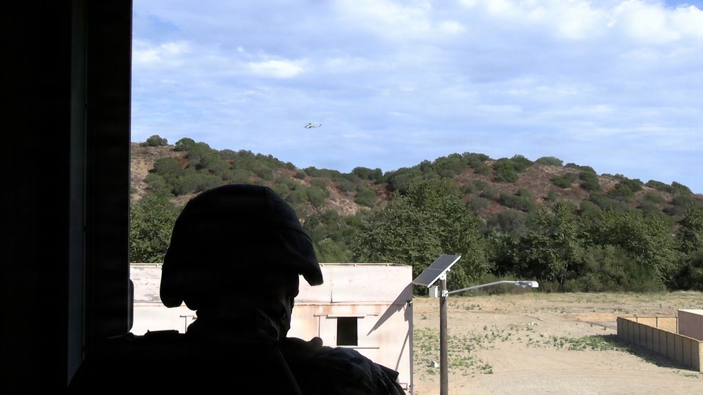 A silhouetted figure wearing a tactical/military helmet is observing from inside a building through an opening. In the background, there is an open training area with low structures, a solar panel on a pole, and a helicopter visible in the sky above a tree-covered hillside. The setting appears to be a military or law enforcement training facility.